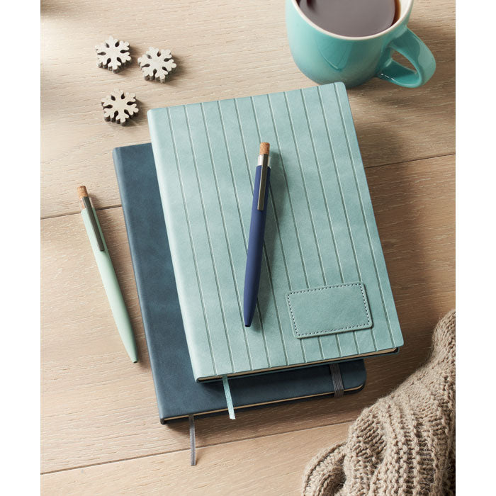 Teal notebook with a pen on a wooden surface with a mug and decorative items.