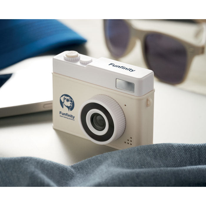 White Funfinity camera on a surface with sunglasses and a blue cap in the background